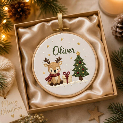 May include: A round wooden embroidery hoop containing white fabric with a hand-stitched design of a brown reindeer wearing a red scarf, a green Christmas tree, a gift box, and gold stars. The name Oliver is embroidered in green script.