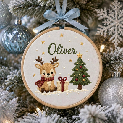 May include: A circular wooden embroidery hoop containing a fabric panel with the embroidered name Oliver in green script. The design features a brown reindeer wearing a red scarf, a decorated green Christmas tree, and a gift box with a red bow.