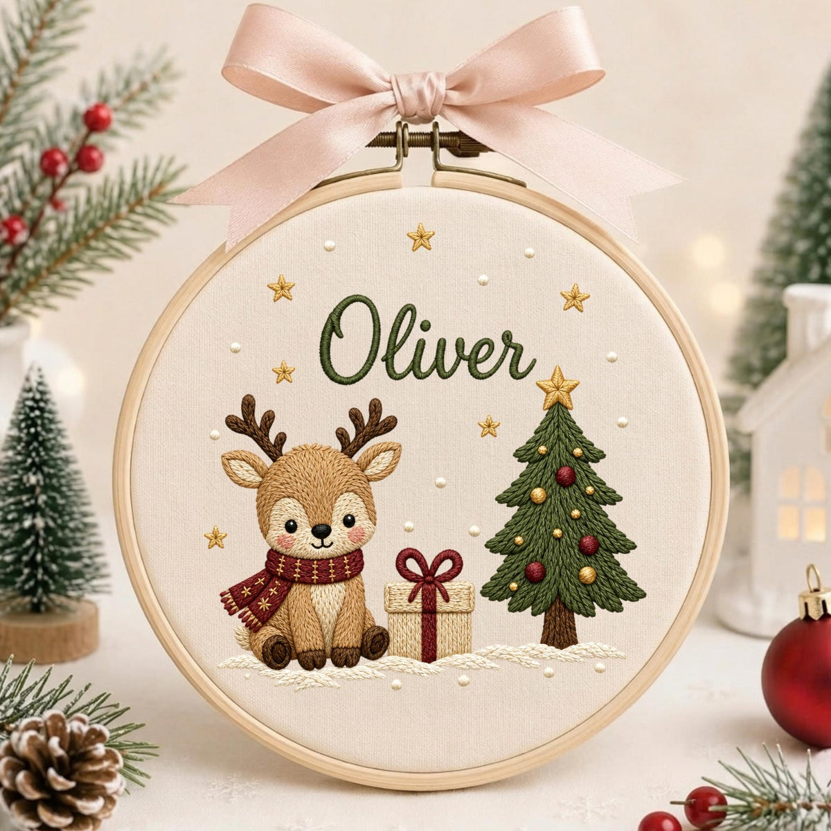 May include: A circular embroidery hoop featuring a cream-colored fabric with a stitched design of a brown reindeer wearing a red scarf, a decorated green Christmas tree, a gift box, and the name Oliver in green, topped with a pink ribbon.