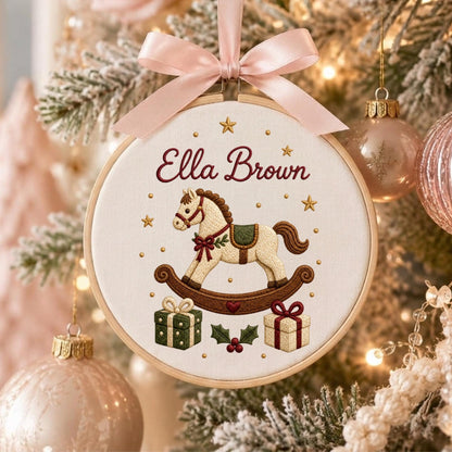May include: A circular embroidery hoop ornament featuring a cream fabric background with the embroidered name Ella Brown in dark red script. A rocking horse, two gift boxes, and small gold stars are stitched in red, green, and gold thread.