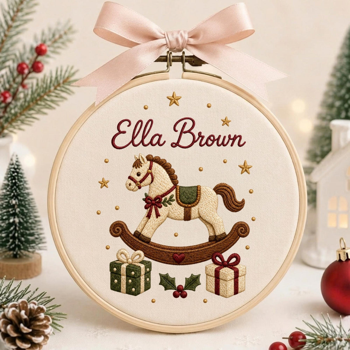 May include: A round wooden embroidery hoop containing cream fabric with a stitched rocking horse, two gift boxes, and holly leaves. The name Ella Brown is embroidered in red script above the horse, topped with a light pink satin ribbon bow.