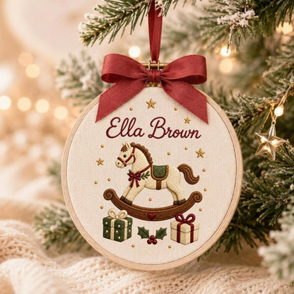 May include: A round wooden embroidery hoop ornament featuring cream fabric with an embroidered rocking horse, two gift boxes, and holly leaves. The name Ella Brown is stitched in red above the horse. A dark red satin ribbon bow is tied at the top.