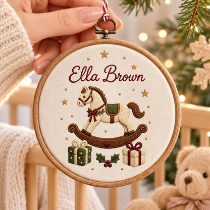 May include: A circular wooden embroidery hoop containing a cream-colored fabric with the embroidered name Ella Brown, a rocking horse, two gift boxes, holly leaves, and small gold stars. The ornament hangs from a red and white striped string.