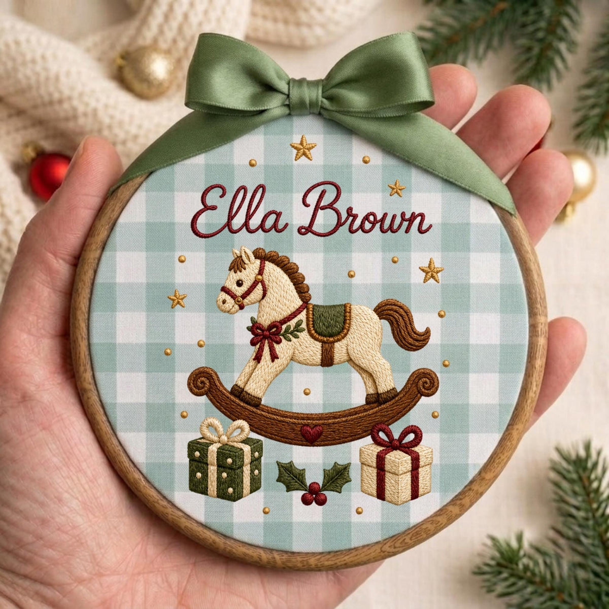 May include: A round wooden embroidery hoop containing fabric with a light blue and white gingham pattern. The fabric features an embroidered rocking horse, two gift boxes, holly leaves, and the name Ella Brown in red script, topped with a green bow.