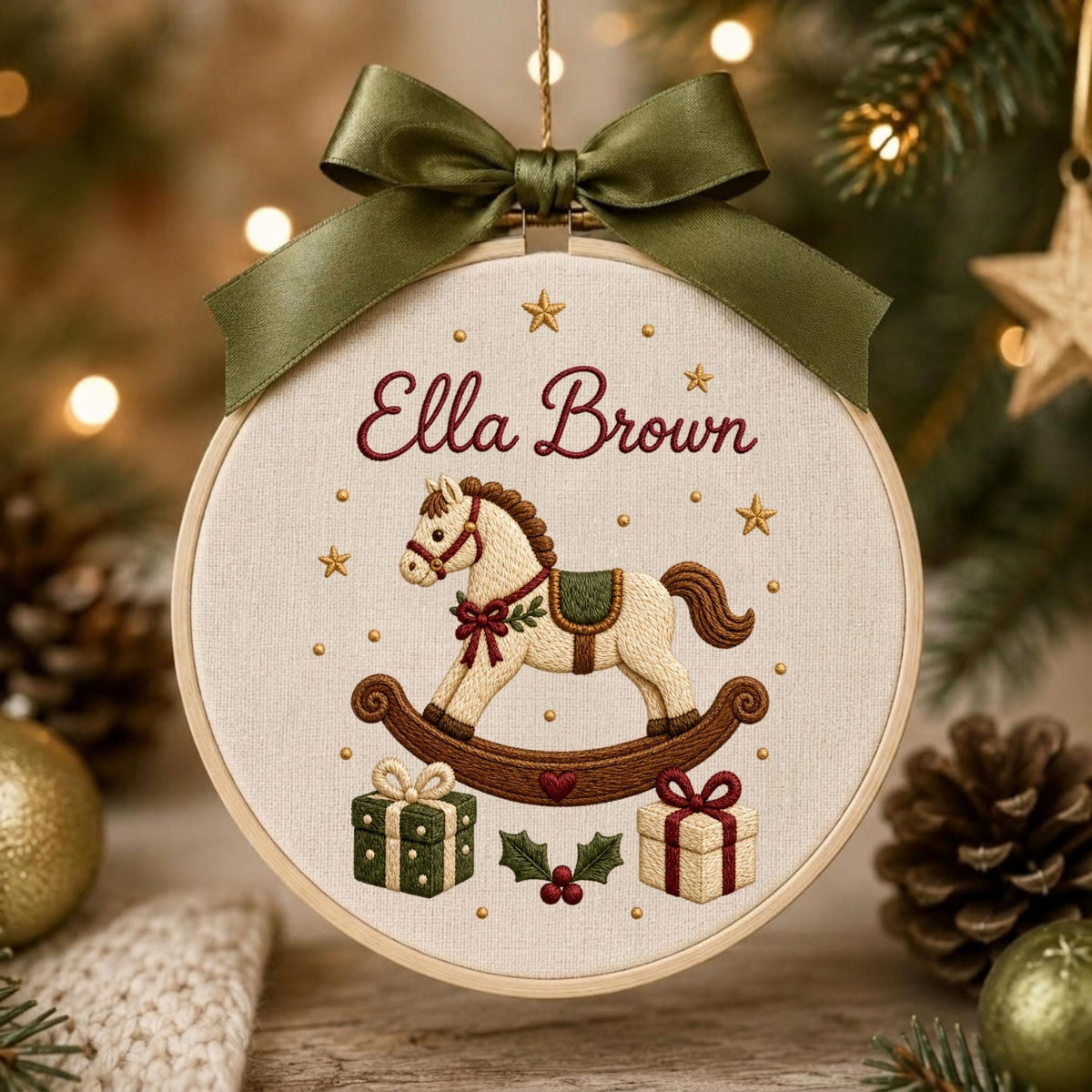May include: A round wooden embroidery hoop ornament featuring a cream-colored fabric background. The embroidery depicts a rocking horse, two gift boxes, and holly leaves. The text Ella Brown is stitched in red. An olive green satin ribbon is tied at the top.