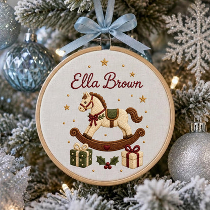 May include: A round wooden embroidery hoop ornament featuring a cream-colored fabric background. The embroidery depicts a rocking horse, two gift boxes, and holly leaves. The text "Ella Brown" is stitched in red above the rocking horse.