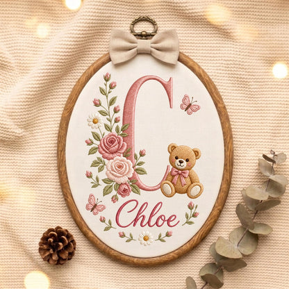 May include: An oval wooden embroidery hoop containing white fabric with a large pink letter C, the name Chloe in pink script, a brown teddy bear with a pink bow, pink roses, green leaves, white daisies, and two small pink butterflies.