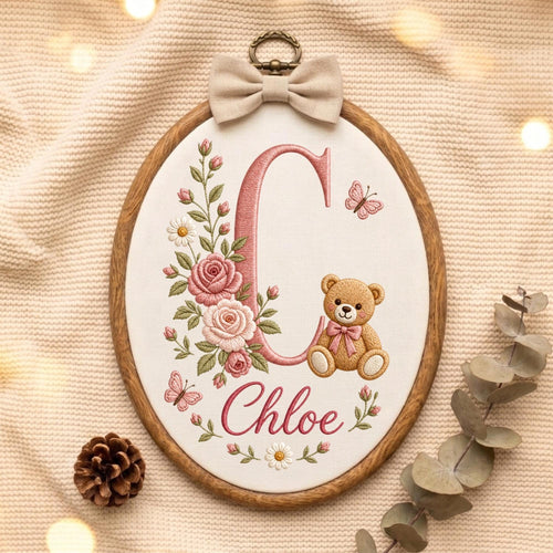 May include: An oval wooden embroidery hoop containing white fabric with a large pink letter C, the name Chloe in pink script, a brown teddy bear with a pink bow, pink roses, green leaves, white daisies, and two small pink butterflies.