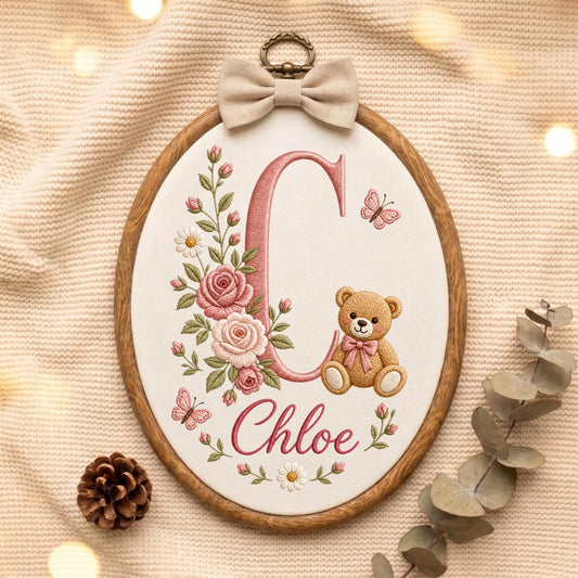 May include: An oval wooden embroidery hoop containing white fabric with a large pink letter C, the name Chloe in pink script, a brown teddy bear with a pink bow, pink roses, green leaves, white daisies, and two small pink butterflies.