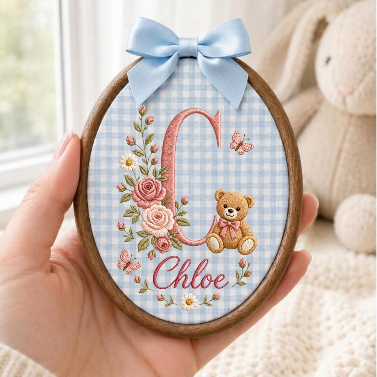 May include: An oval wooden embroidery hoop containing fabric with a light blue gingham pattern. The design features a large pink letter C, a brown teddy bear, pink roses, daisies, and small butterflies. The name Chloe is embroidered in pink.