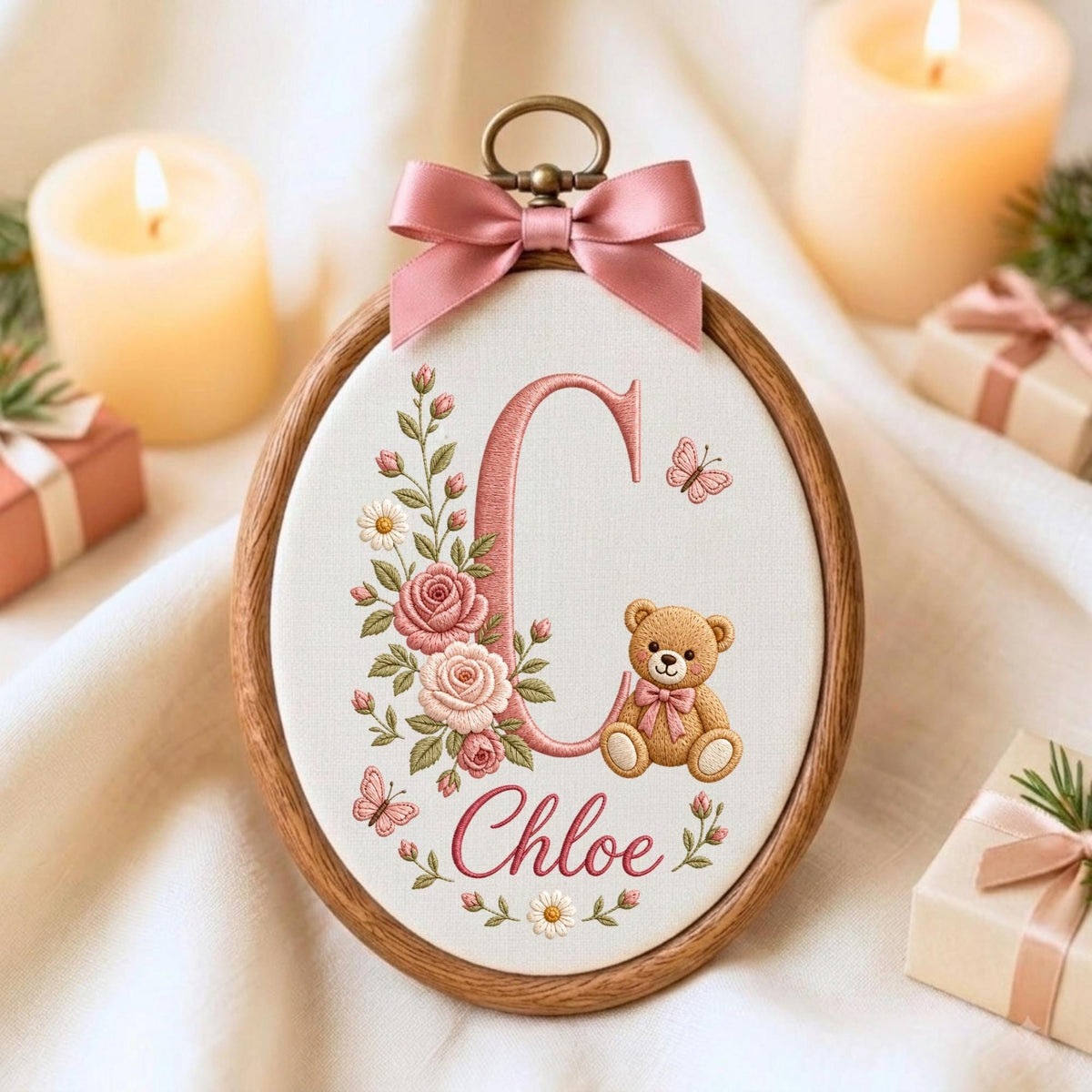 May include: An oval wooden embroidery hoop containing white fabric with a stitched design featuring a large pink letter C, a teddy bear, pink roses, daisies, and butterflies. The name Chloe is embroidered below, topped with a pink satin bow.