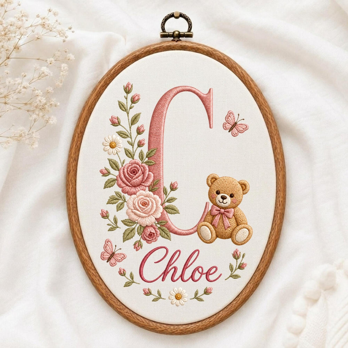 May include: An oval wooden embroidery hoop containing white fabric with a stitched design. The design features a large pink letter C, a brown teddy bear with a pink bow, pink roses, green leaves, white daisies, and two small pink butterflies.