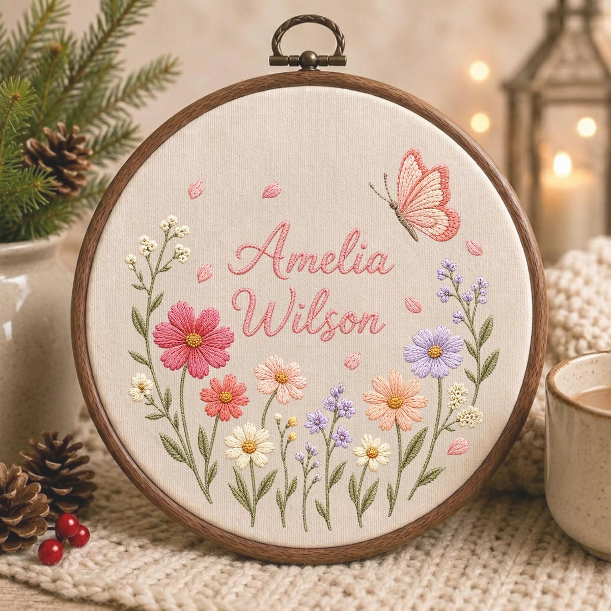 May include: A circular wooden embroidery hoop containing cream-colored fabric with the embroidered name Amelia Wilson in pink script. The design features a colorful floral arrangement of pink, peach, white, and purple flowers with a pink butterfly.