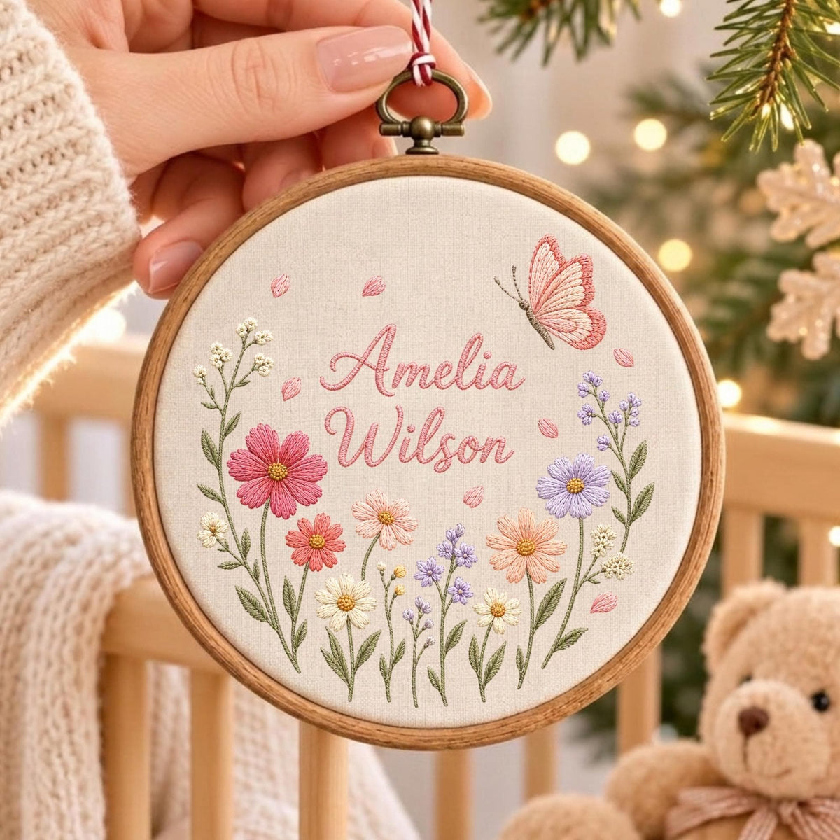 May include: A circular wooden embroidery hoop containing off-white fabric with the embroidered name Amelia Wilson in pink script. The design features a pink butterfly and a colorful arrangement of embroidered wildflowers in shades of pink, purple, and white.