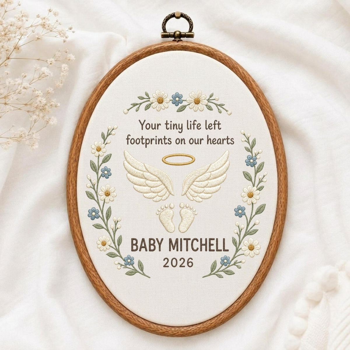 May include: An oval wooden embroidery hoop containing white fabric with floral embroidery. The text reads: Your tiny life left footprints on our hearts, BABY MITCHELL, 2026. It features embroidered angel wings, a halo, and baby footprints in cream.