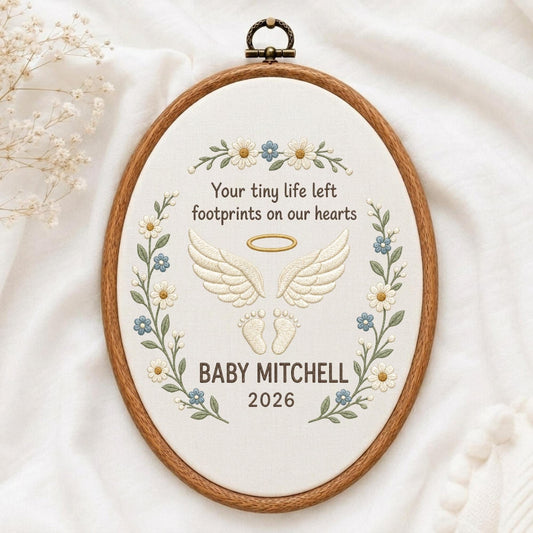 May include: An oval wooden embroidery hoop containing white fabric with floral embroidery. The text reads: Your tiny life left footprints on our hearts, BABY MITCHELL, 2026. It features embroidered angel wings, a halo, and baby footprints in cream.