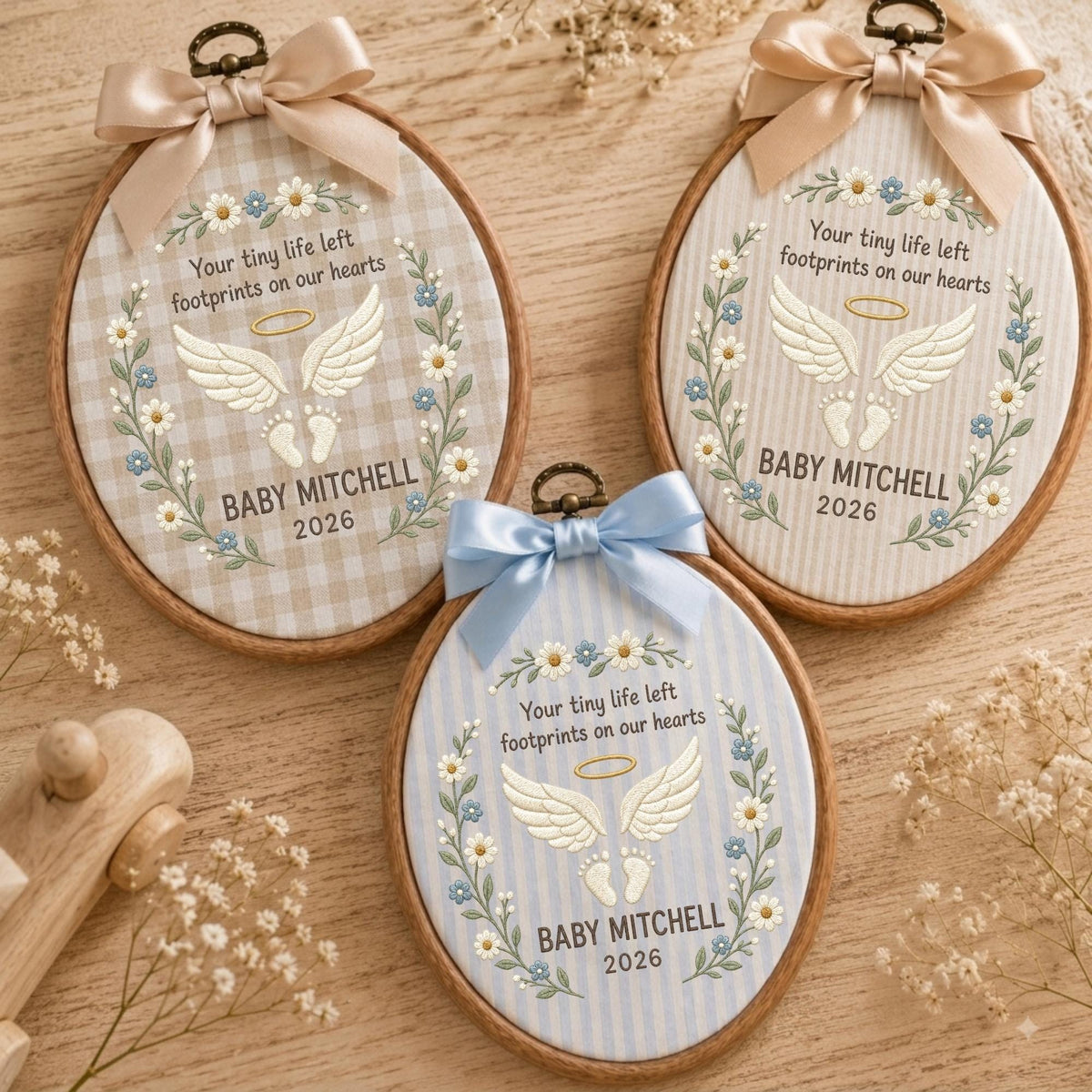 May include: Three oval wooden embroidery hoops featuring fabric backgrounds with gingham or striped patterns. Each hoop displays embroidered angel wings, a halo, baby footprints, and the text Your tiny life left footprints on our hearts, BABY MITCHELL, 2026, framed by floral wreaths and topped with satin bows.