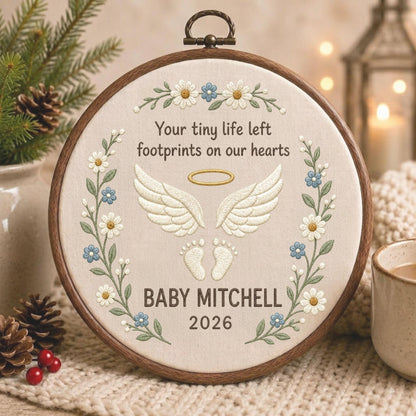 May include: A circular wooden embroidery hoop containing cream-colored fabric with stitched text reading Your tiny life left footprints on our hearts, BABY MITCHELL, and 2026. The design features embroidered white angel wings, a gold halo, and baby footprints.
