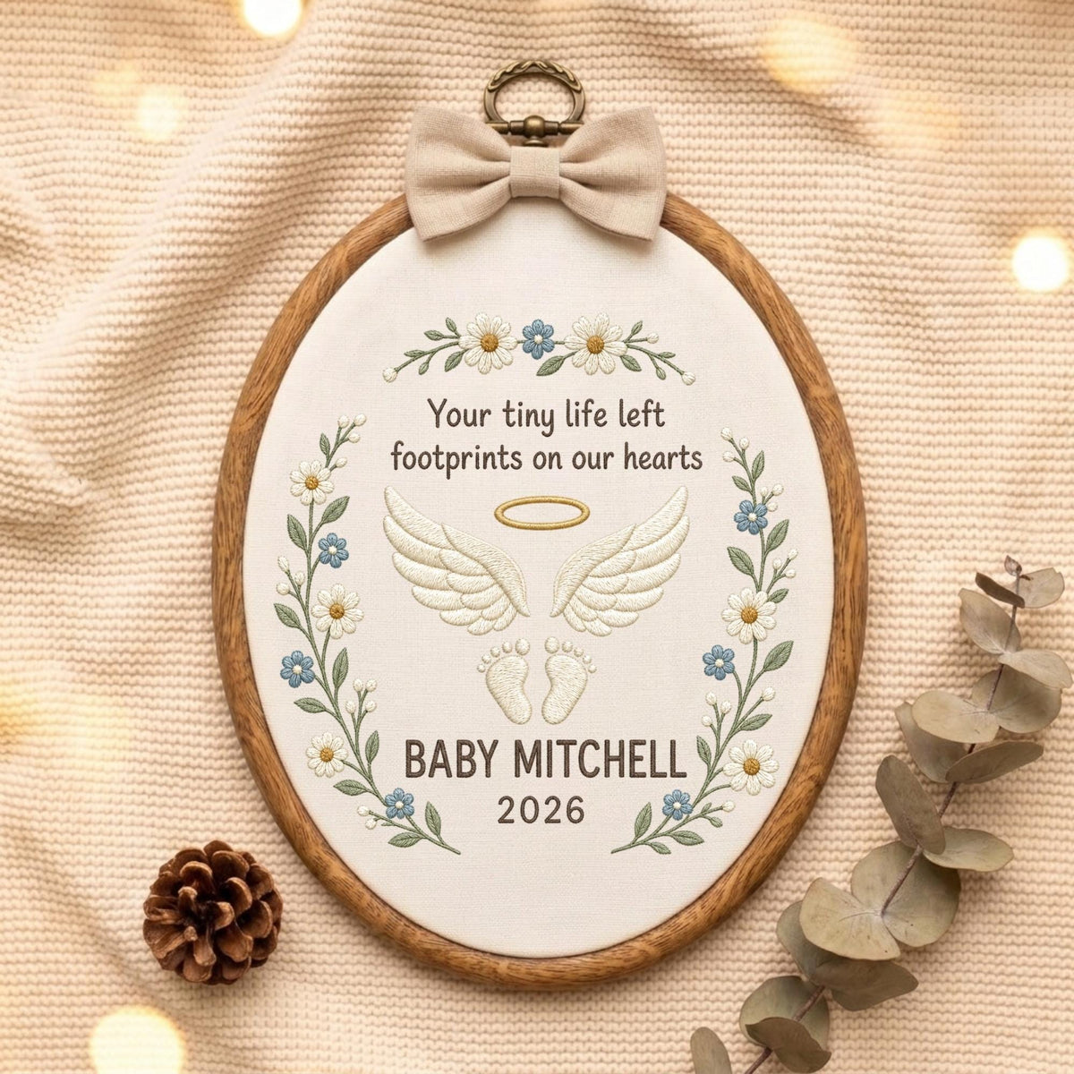 May include: An oval embroidery hoop featuring a beige fabric background with embroidered white angel wings, a gold halo, and baby footprints. The text reads: Your tiny life left footprints on our hearts, BABY MITCHELL, 2026. A beige bow sits at the top.