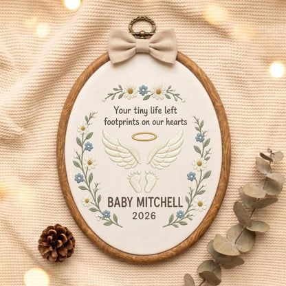 May include: An oval embroidery hoop featuring a beige fabric background with embroidered white angel wings, a gold halo, and baby footprints. The text reads: Your tiny life left footprints on our hearts, BABY MITCHELL, 2026. A beige bow sits at the top.