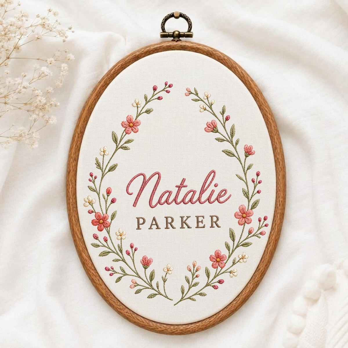 May include: An oval wooden embroidery hoop containing white fabric with hand-stitched floral embroidery. The design features a wreath of pink and white flowers with green leaves surrounding the embroidered text Natalie PARKER in pink and brown.