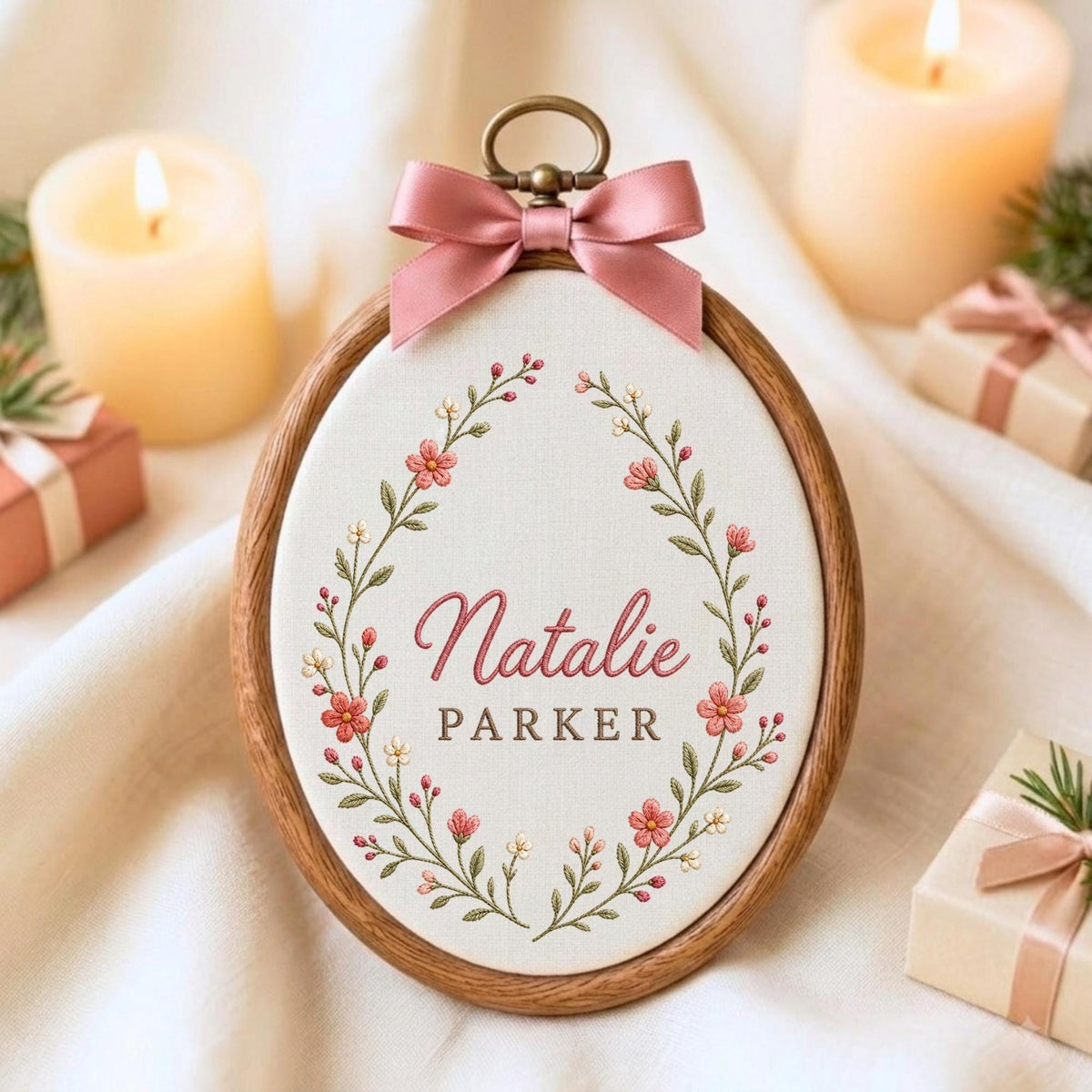 May include: An oval wooden embroidery hoop frame containing white fabric with an embroidered floral wreath in pink and green. The name Natalie Parker is stitched in the center in pink and brown. A dusty pink satin ribbon bow sits at the top.