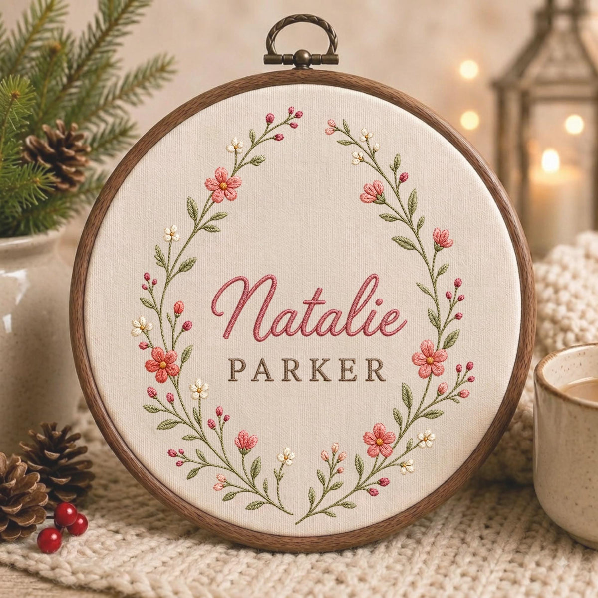May include: A circular wooden embroidery hoop containing off-white fabric with a hand-stitched floral wreath in shades of pink, white, and green. The name Natalie is embroidered in pink script above the name PARKER in brown block letters.