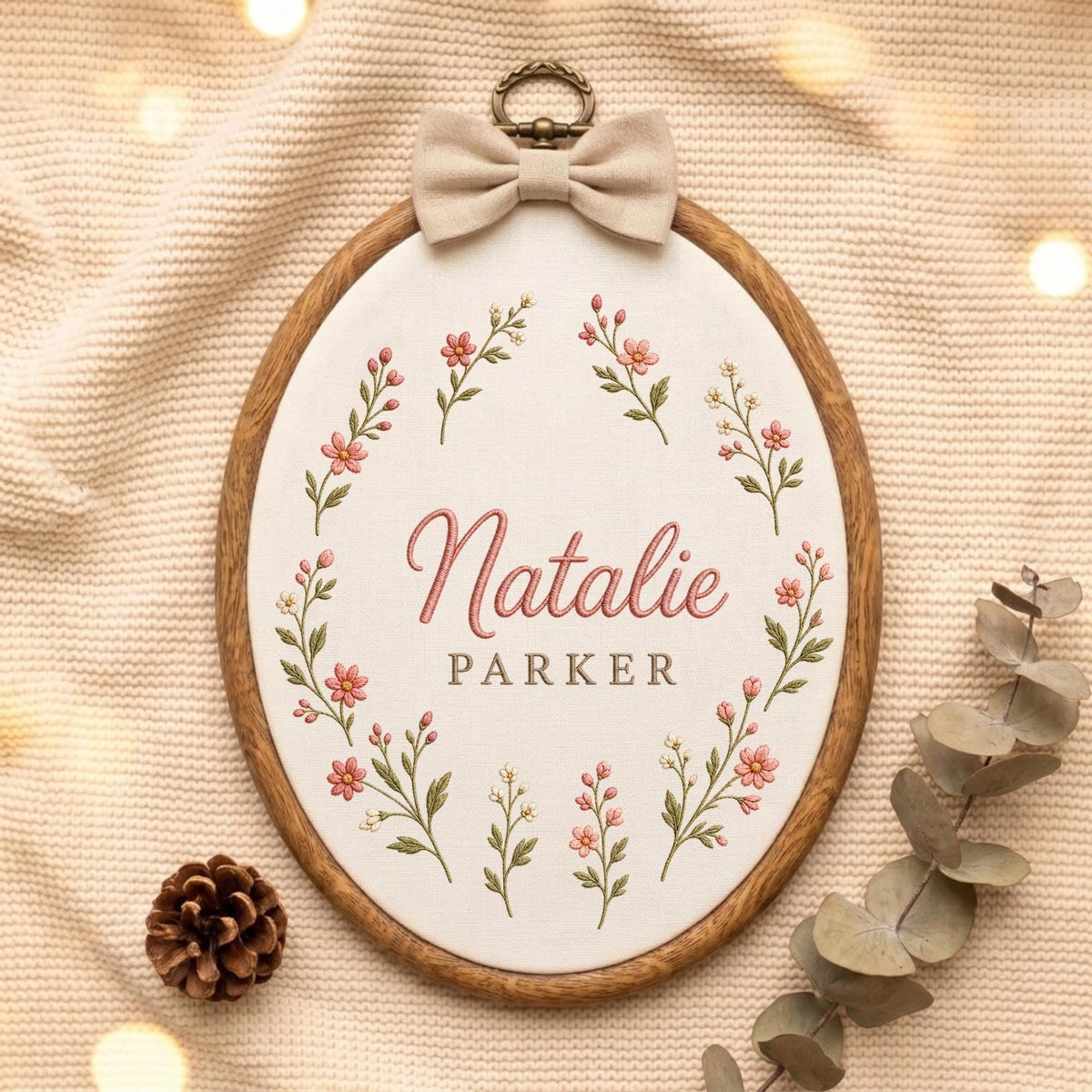 May include: An oval wooden embroidery hoop containing white fabric with the embroidered name Natalie Parker in pink and brown script and block lettering, surrounded by a floral wreath of pink flowers and green leaves, topped with a beige bow.