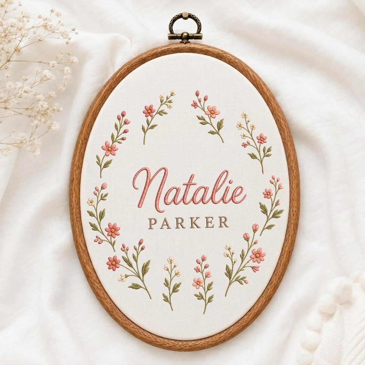 May include: An oval wooden embroidery hoop containing white fabric with a floral wreath design in pink and green thread. The name Natalie is embroidered in pink script, with the name Parker in brown block letters centered within the floral frame.