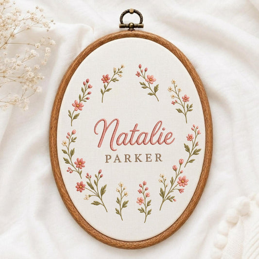 May include: An oval wooden embroidery hoop containing white fabric with a floral wreath design in pink and green thread. The name Natalie is embroidered in pink script, with the name Parker in brown block letters centered within the floral frame.