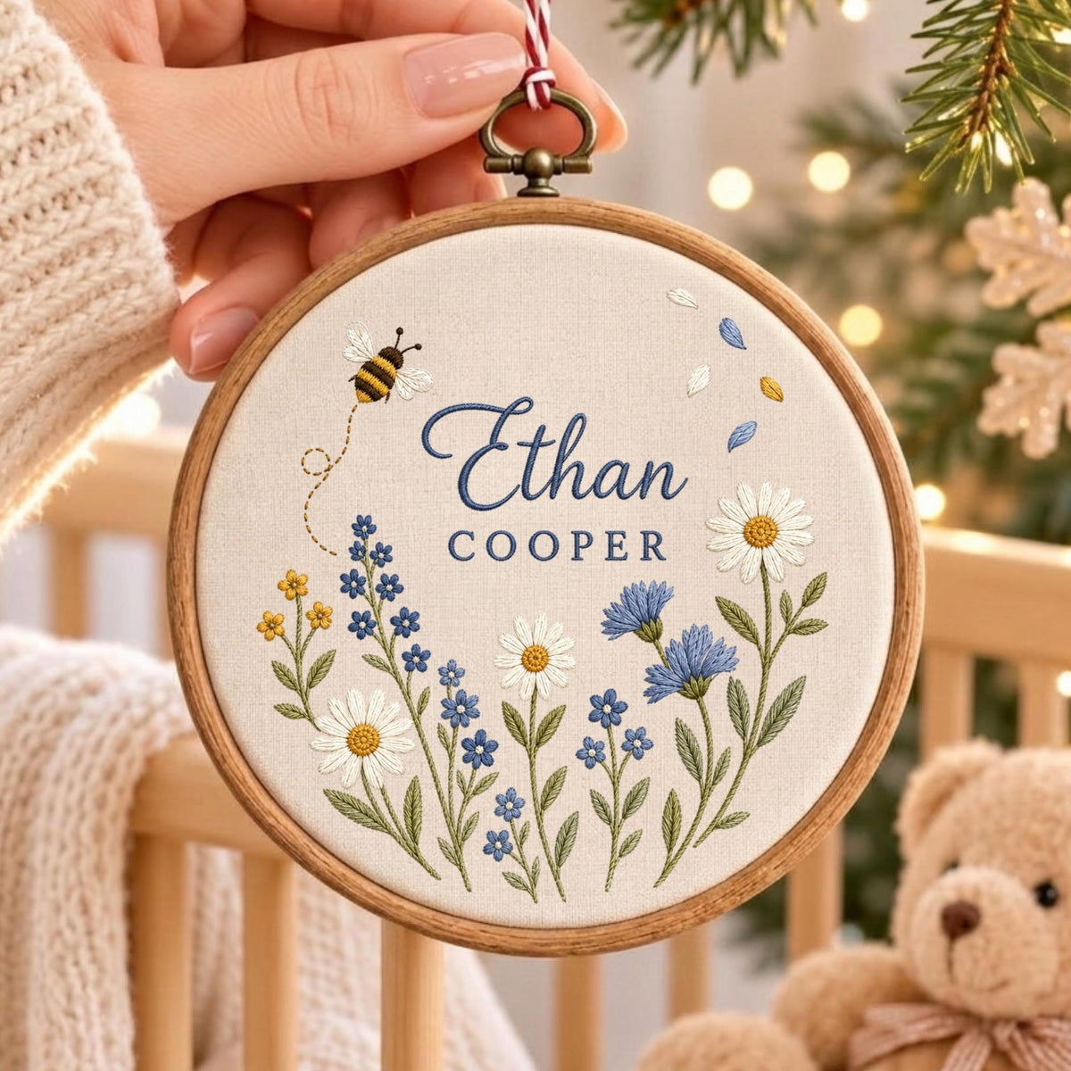 May include: A circular wooden embroidery hoop containing off-white fabric with the embroidered name Ethan Cooper in blue script and block letters. The design features a small bee, white daisies, and blue wildflowers with green stems and leaves.