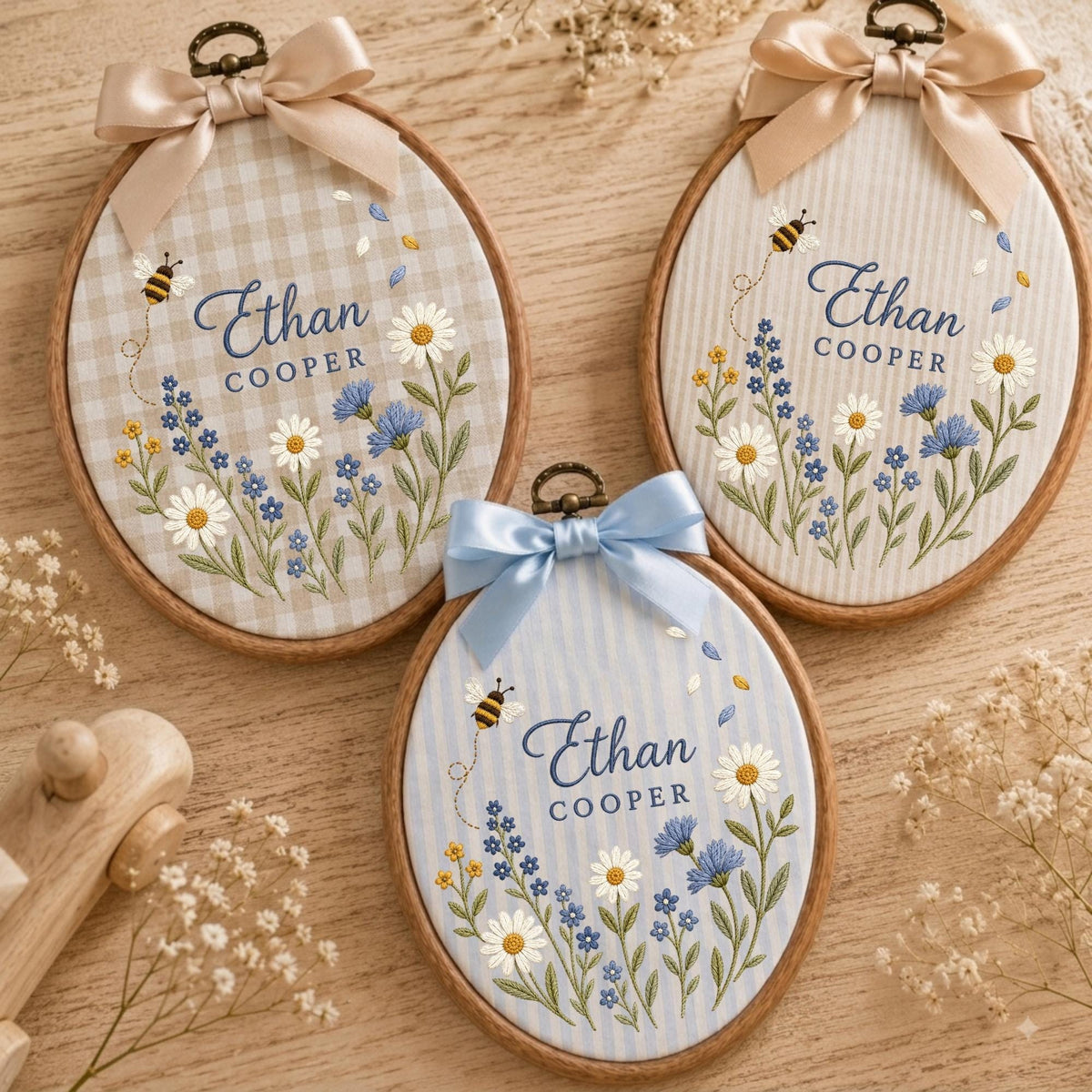 May include: Three oval wooden embroidery hoops featuring fabric backgrounds with gingham or striped patterns. Each hoop displays embroidered wildflowers, a small bee, and the text Ethan Cooper in blue script, topped with a satin ribbon bow.