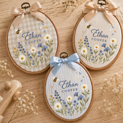 May include: Three oval wooden embroidery hoops featuring fabric backgrounds with gingham or striped patterns. Each hoop displays embroidered wildflowers, a small bee, and the text Ethan Cooper in blue script, topped with a satin ribbon bow.