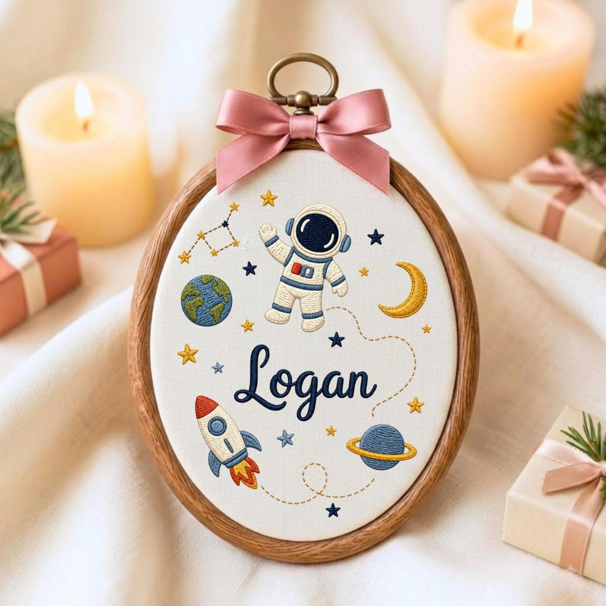 May include: An oval embroidery hoop frame containing white fabric with a space-themed design. The embroidery features an astronaut, a rocket, planets, stars, and the name Logan in dark blue script. A pink satin bow is attached to the top.