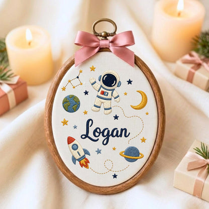 May include: An oval embroidery hoop frame containing white fabric with a space-themed design. The embroidery features an astronaut, a rocket, planets, stars, and the name Logan in dark blue script. A pink satin bow is attached to the top.