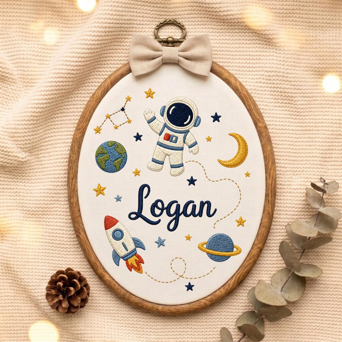 May include: An oval embroidery hoop featuring a white fabric background with a space-themed design. The embroidery includes an astronaut, a rocket, planets, stars, and the name Logan in dark blue script. A beige fabric bow sits at the top.