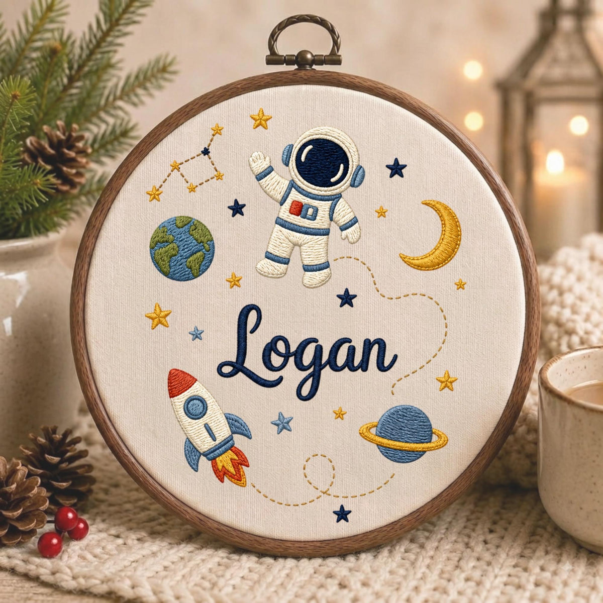 May include: A circular wooden embroidery hoop containing off-white fabric with a space-themed design. The embroidery features a white astronaut, a rocket ship, the Earth, a planet with rings, a crescent moon, stars, and the name Logan in blue.