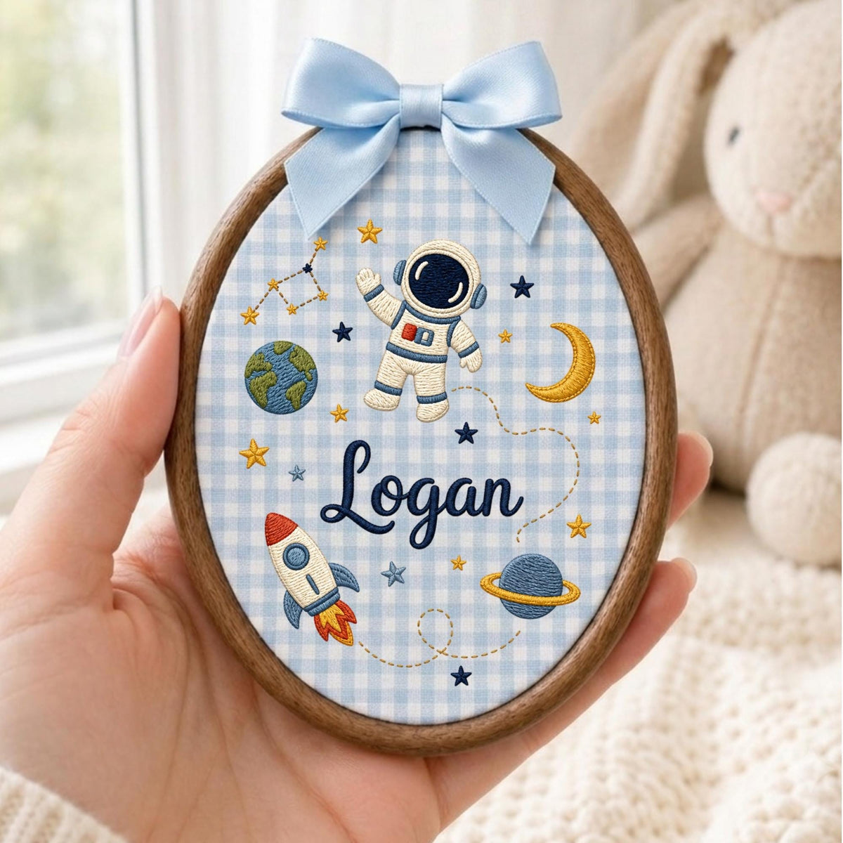 May include: An oval embroidery hoop featuring a light blue and white gingham fabric background. The design includes an embroidered astronaut, a rocket, planets, stars, and the name Logan in dark blue script, topped with a light blue satin bow.