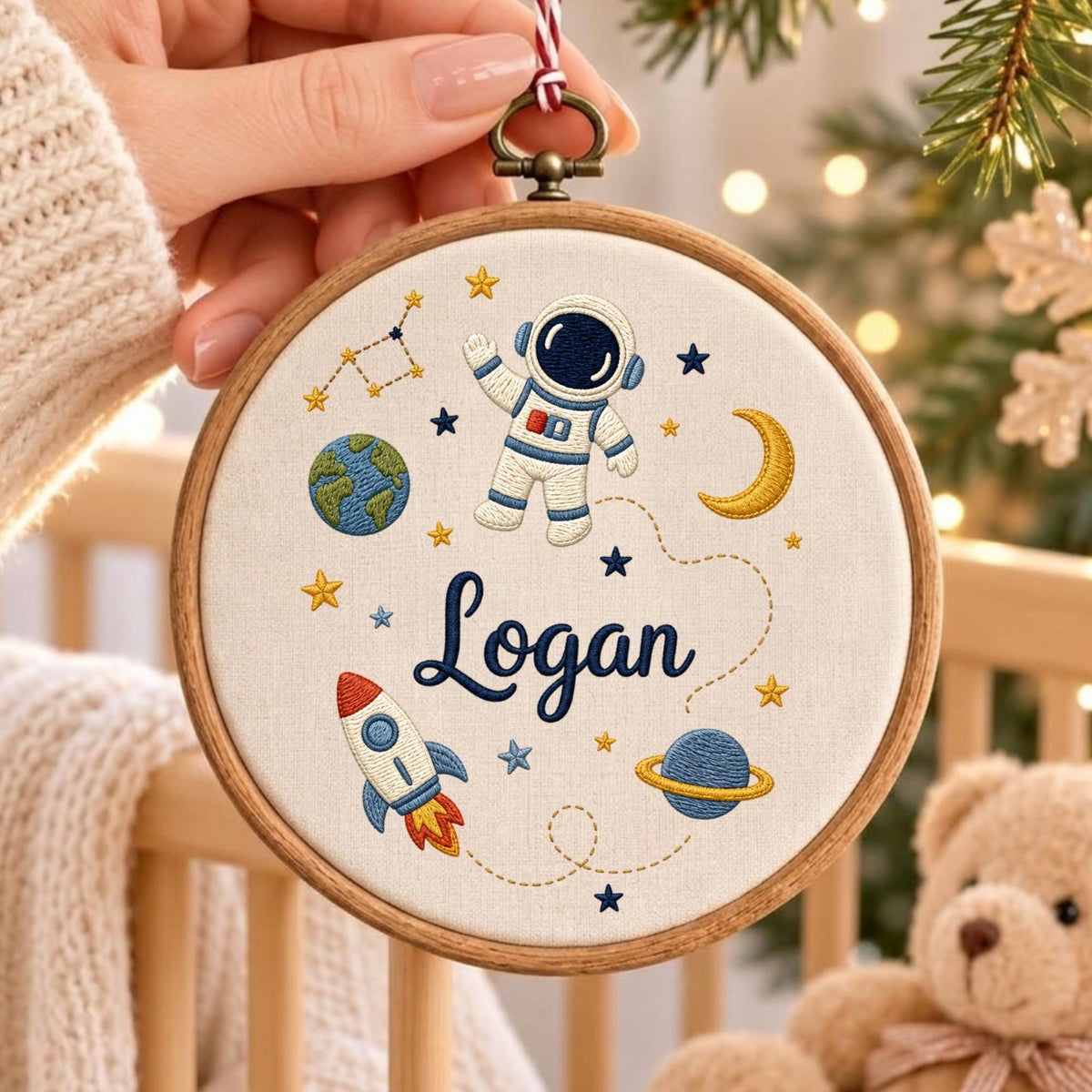 May include: A circular wooden embroidery hoop containing off-white fabric with space-themed embroidery. The design features an astronaut, a rocket, planets, stars, and a crescent moon. The name Logan is embroidered in dark blue script in the center.