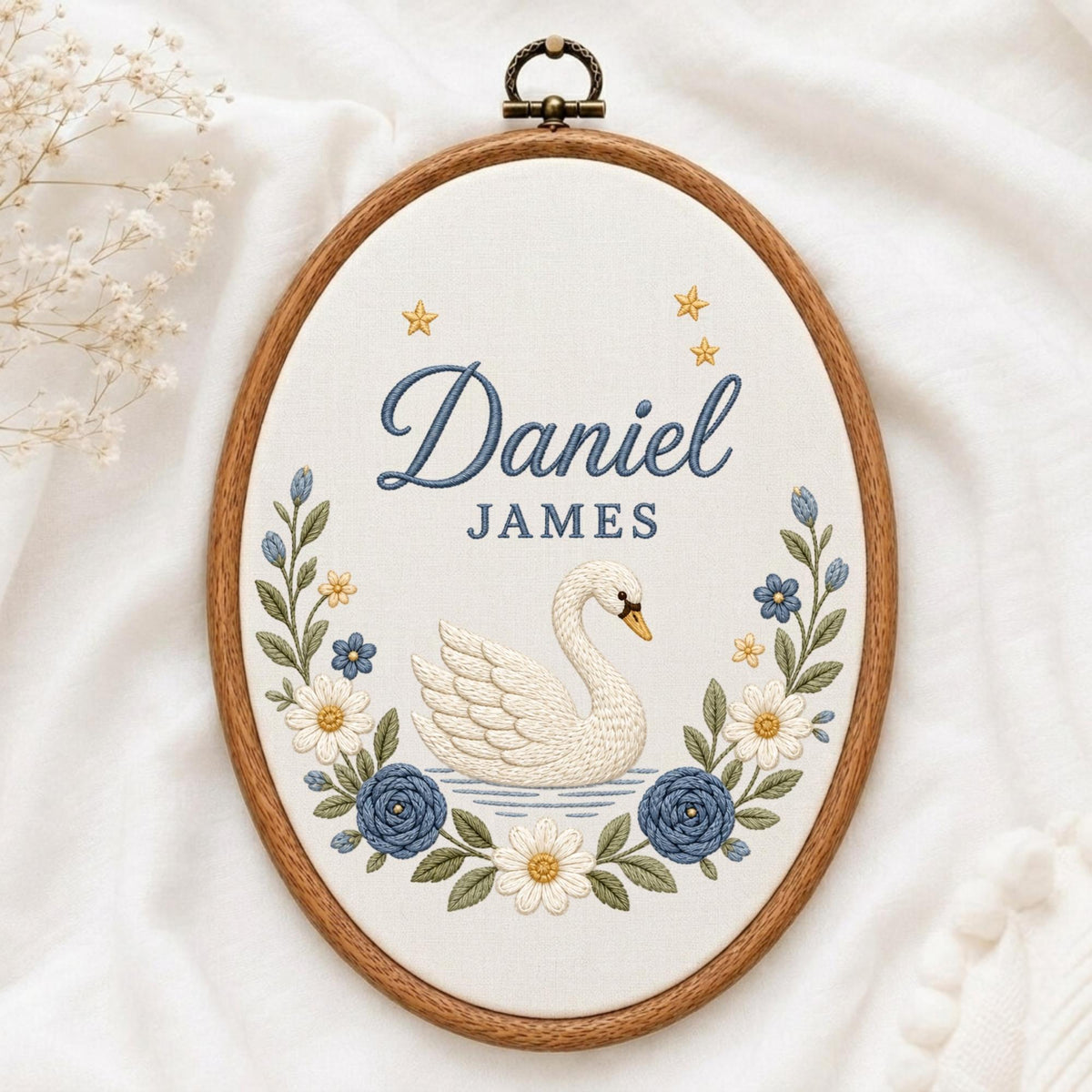 May include: An oval wooden embroidery hoop containing white fabric with a hand-stitched design. The embroidery features a white swan, blue and white flowers, green leaves, and yellow stars. The text Daniel JAMES is stitched in blue thread.