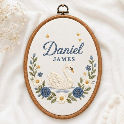 May include: An oval wooden embroidery hoop containing white fabric with a hand-stitched design. The embroidery features a white swan, blue and white flowers, green leaves, and yellow stars. The text Daniel JAMES is stitched in blue thread.