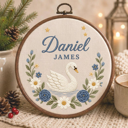May include: A circular wooden embroidery hoop containing a cream-colored fabric panel. The embroidery features a white swan centered above blue water, surrounded by a floral wreath of blue roses and white daisies. The name Daniel James is stitched in blue.