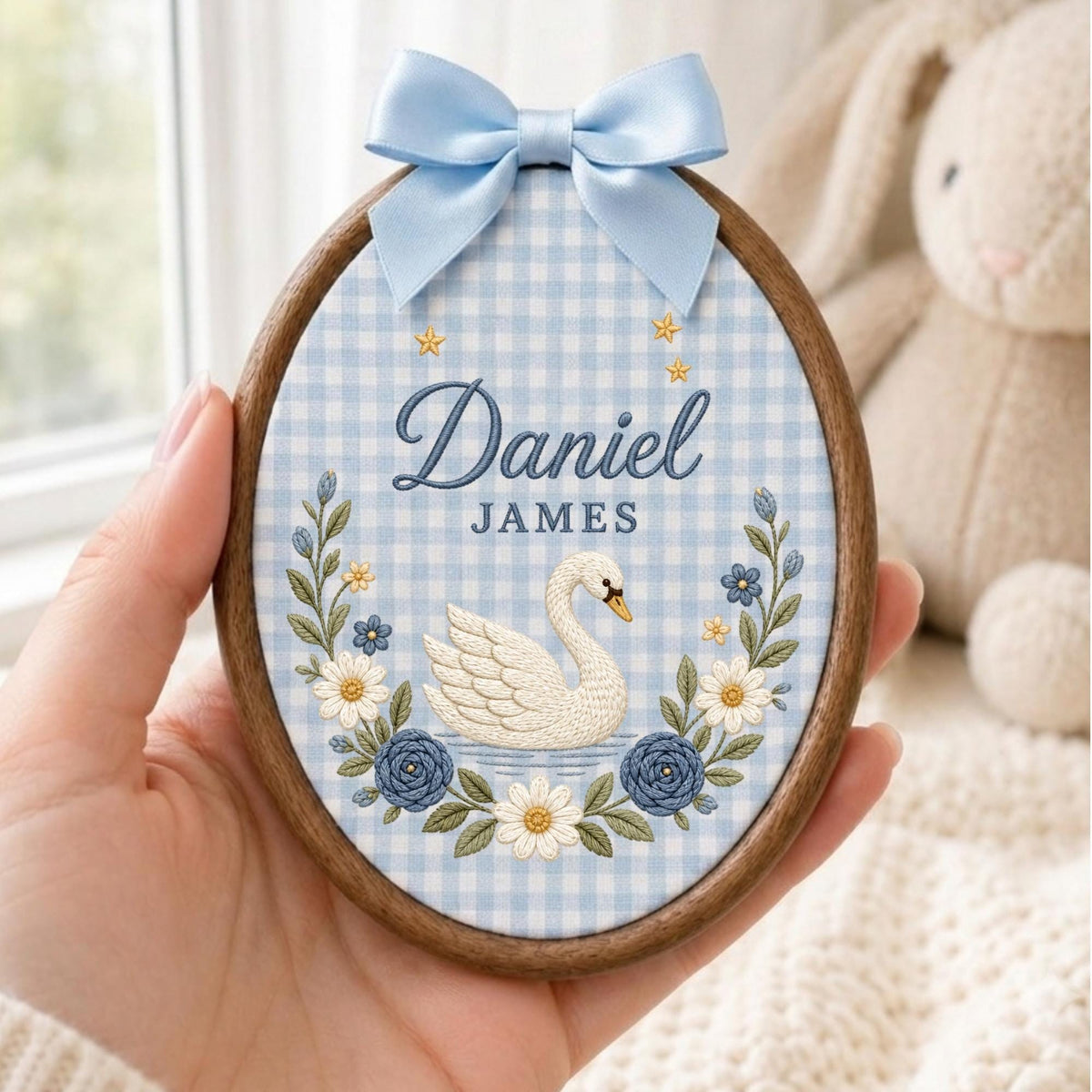 May include: An oval wooden embroidery hoop containing fabric with a light blue and white gingham pattern. The embroidery features a white swan, blue and white flowers, and the text "Daniel JAMES" in blue script and block lettering with a blue bow.
