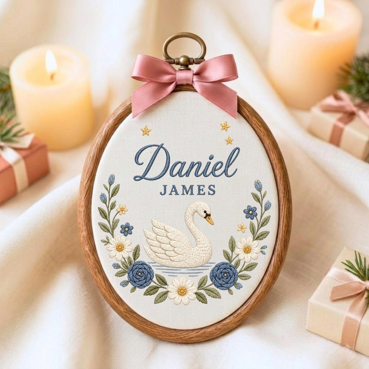 May include: An oval embroidery hoop featuring a white fabric background with a centered white swan illustration, blue floral accents, and the text Daniel JAMES in blue. A pink satin ribbon bow is attached to the top metal hanging loop.