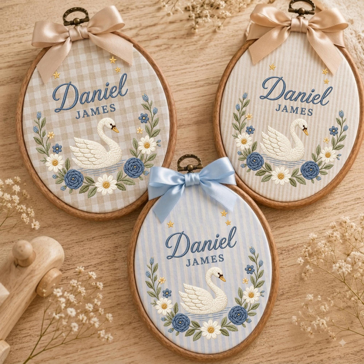 May include: Three oval embroidery hoops featuring the name Daniel James in blue script, a white swan, and a floral wreath. Each hoop is topped with a satin ribbon bow and displays a different fabric background: beige gingham, beige stripes, and blue stripes.