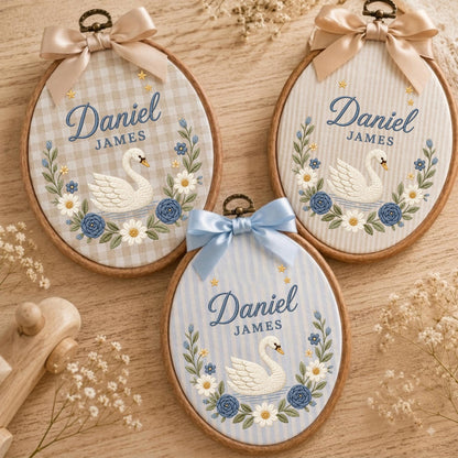 May include: Three oval embroidery hoops featuring the name Daniel James in blue script, a white swan, and a floral wreath. Each hoop is topped with a satin ribbon bow and displays a different fabric background: beige gingham, beige stripes, and blue stripes.