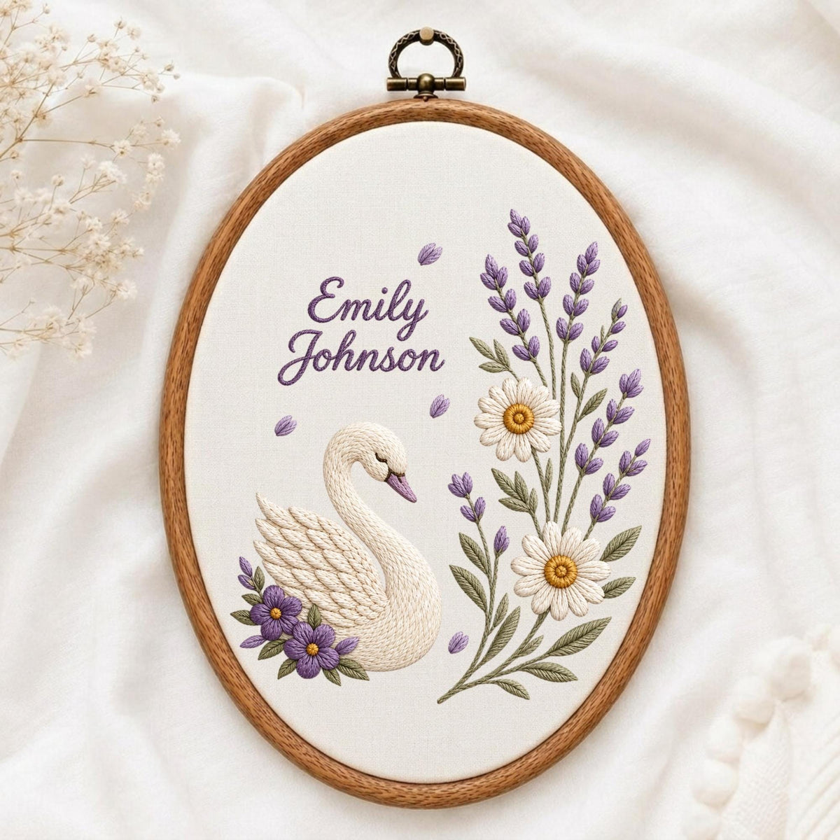 May include: An oval embroidery hoop containing white fabric with a hand-stitched design of a white swan, purple flowers, lavender sprigs, and white daisies. The text "Emily Johnson" is embroidered in purple script above the swan.