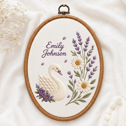 May include: An oval embroidery hoop containing white fabric with a hand-stitched design of a white swan, purple flowers, lavender sprigs, and white daisies. The text "Emily Johnson" is embroidered in purple script above the swan.