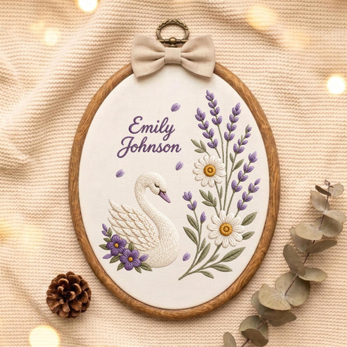May include: An oval embroidery hoop featuring a white fabric background with a stitched white swan, purple flowers, and lavender sprigs. The text "Emily Johnson" is embroidered in purple. A beige fabric bow sits atop the wooden frame.