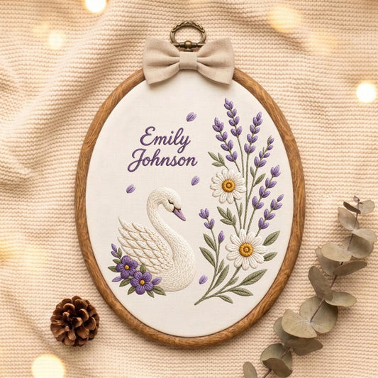 May include: An oval embroidery hoop featuring a white fabric background with a stitched white swan, purple flowers, and lavender sprigs. The text "Emily Johnson" is embroidered in purple. A beige fabric bow sits atop the wooden frame.